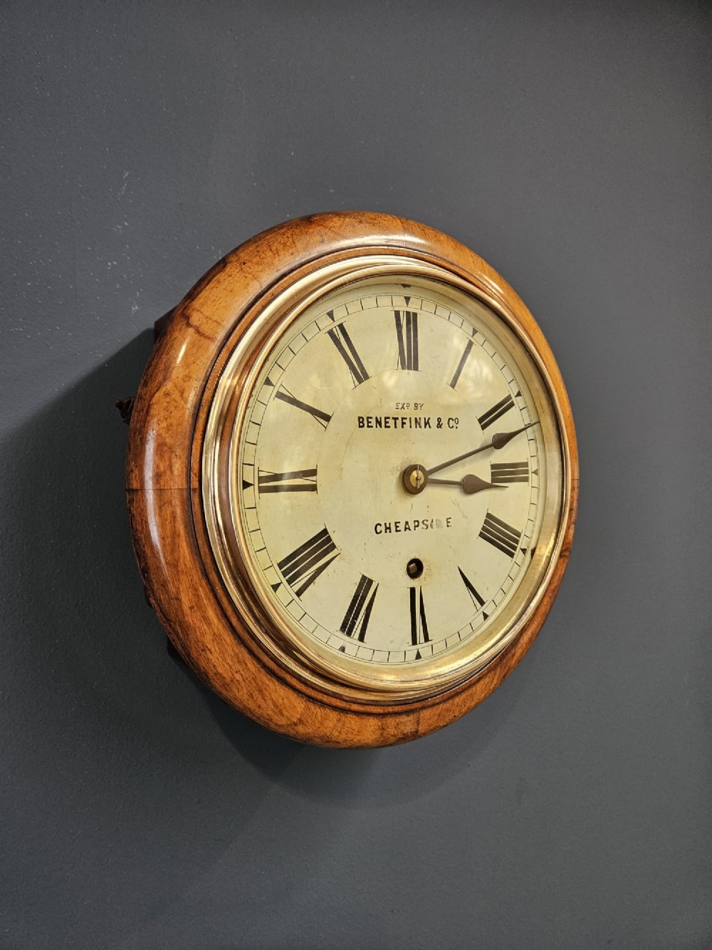 A circular Victorian wall clock featuring a turned warm brown walnut frame, brass-rimmed glass door, and off-white dial marked with Roman numerals and Benetfink and Co Cheapside. View III