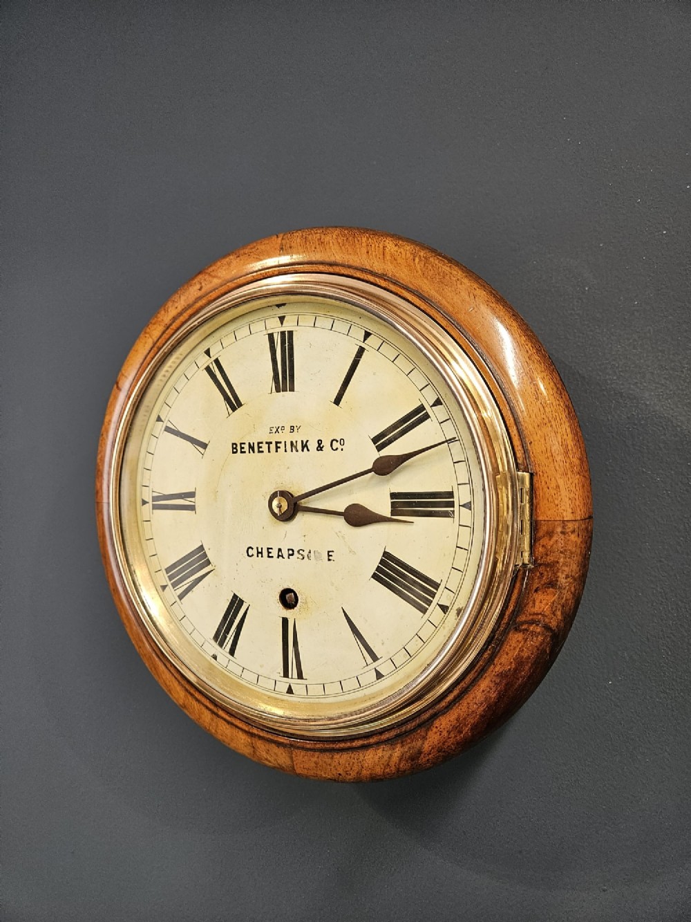 A circular Victorian wall clock featuring a turned warm brown walnut frame, brass-rimmed glass door, and off-white dial marked with Roman numerals and Benetfink and Co Cheapside. View IV
