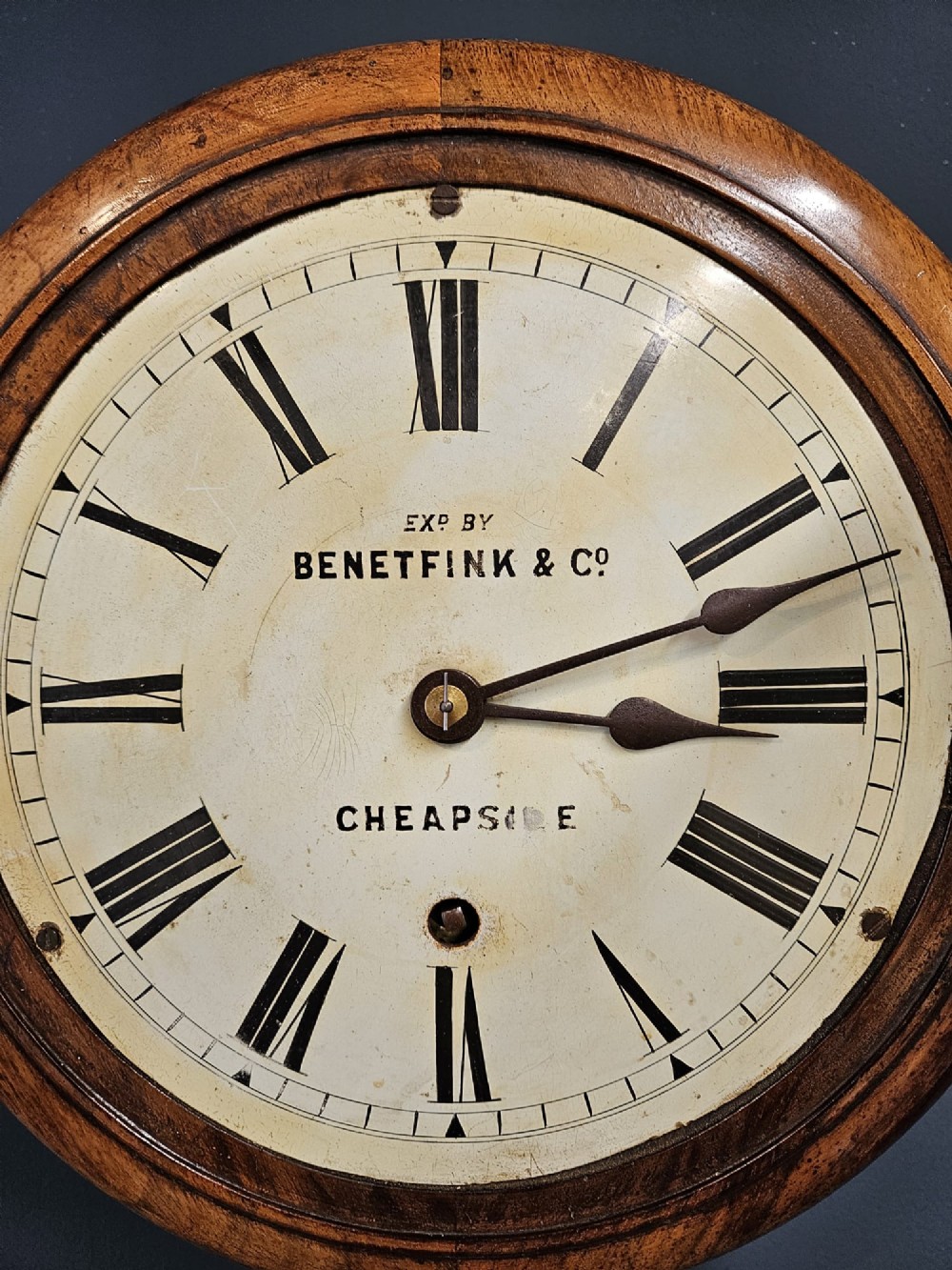 A circular Victorian wall clock featuring a turned warm brown walnut frame, brass-rimmed glass door, and off-white dial marked with Roman numerals and Benetfink and Co Cheapside. View V