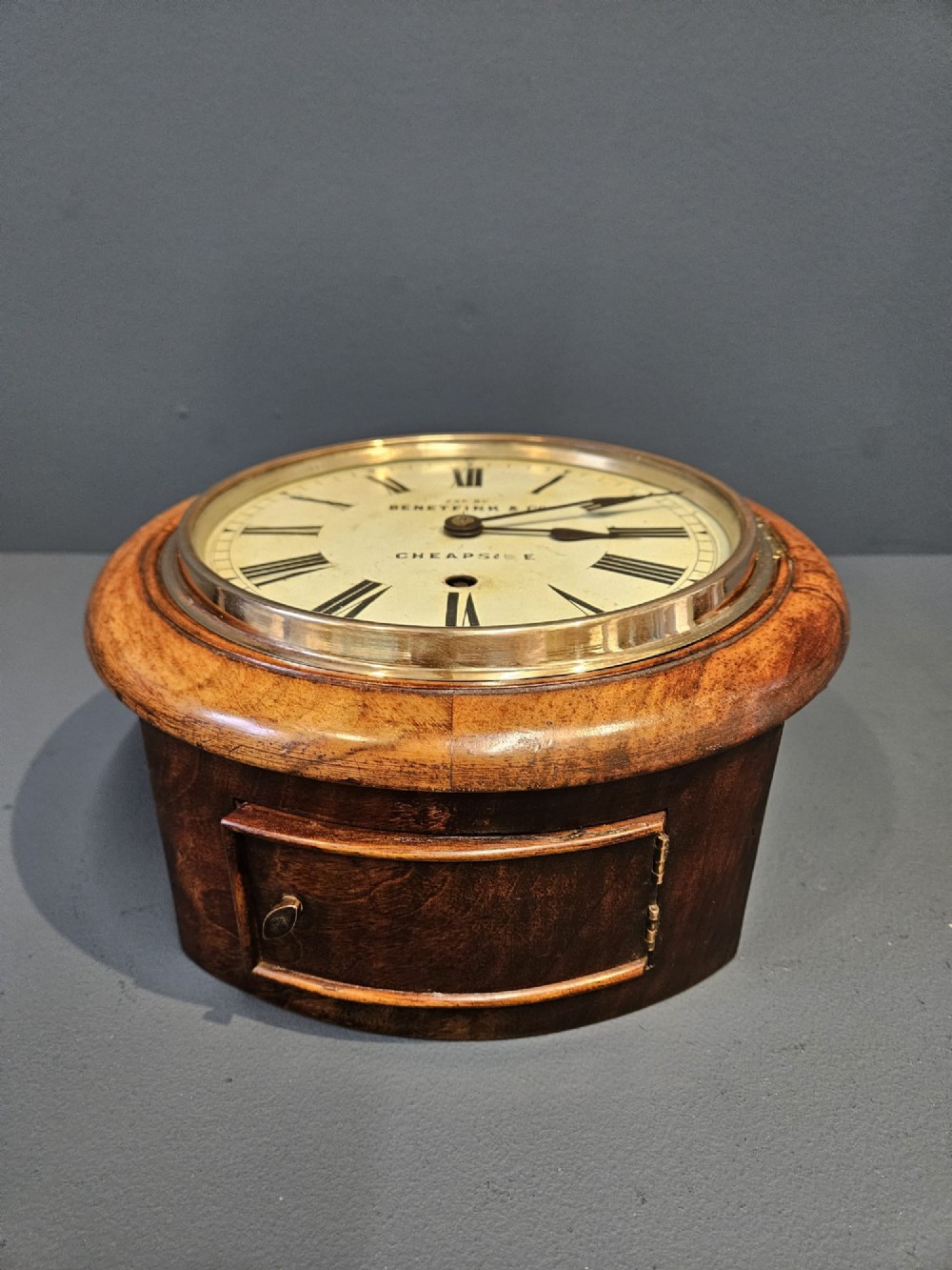 A circular Victorian wall clock featuring a turned warm brown walnut frame, brass-rimmed glass door, and off-white dial marked with Roman numerals and Benetfink and Co Cheapside. View VII