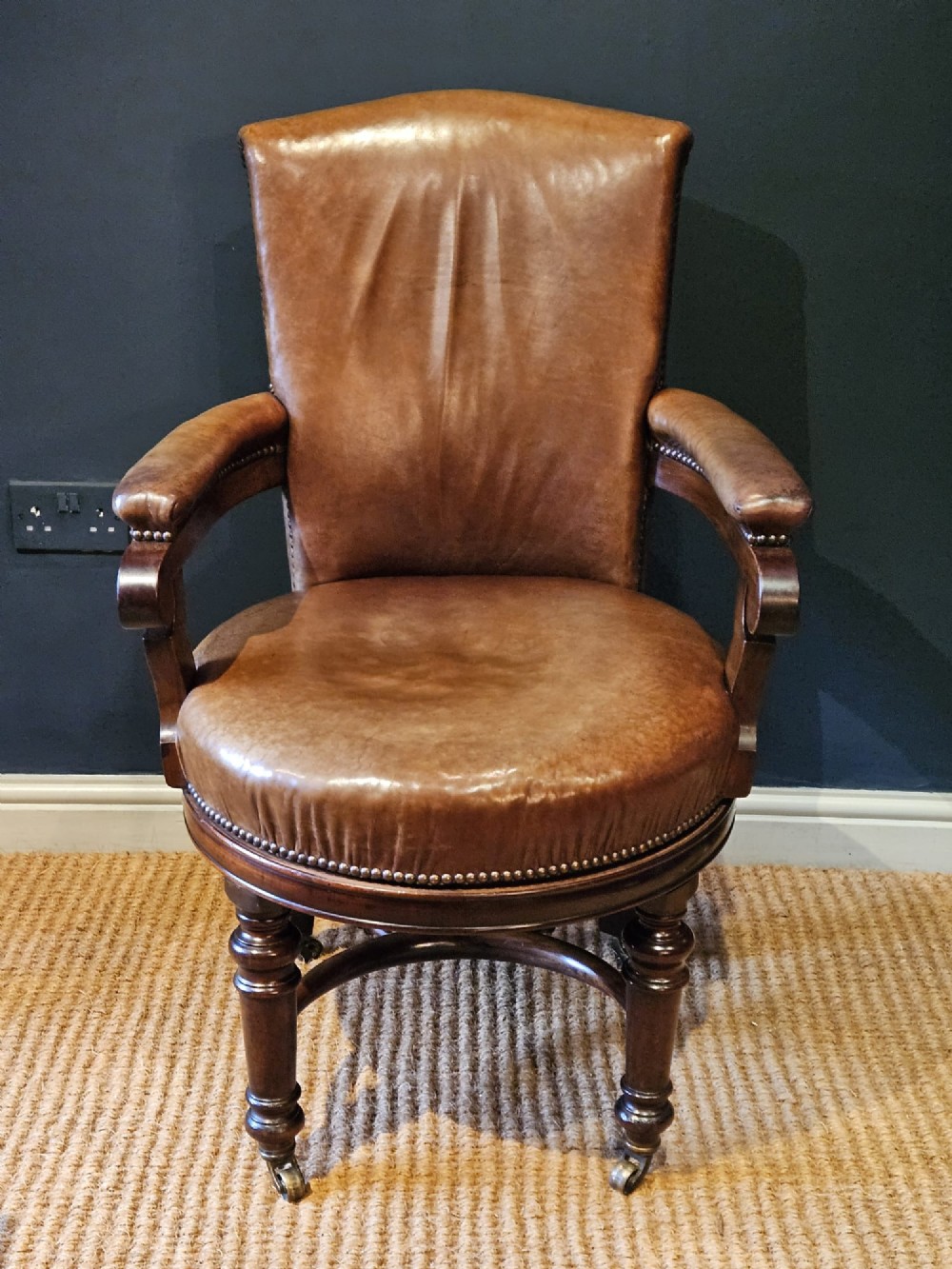 A Victorian revolving desk chair upholstered in tan hide leather with brass stud trim, curved armrests, and turned mahogany legs on brass castors. View II