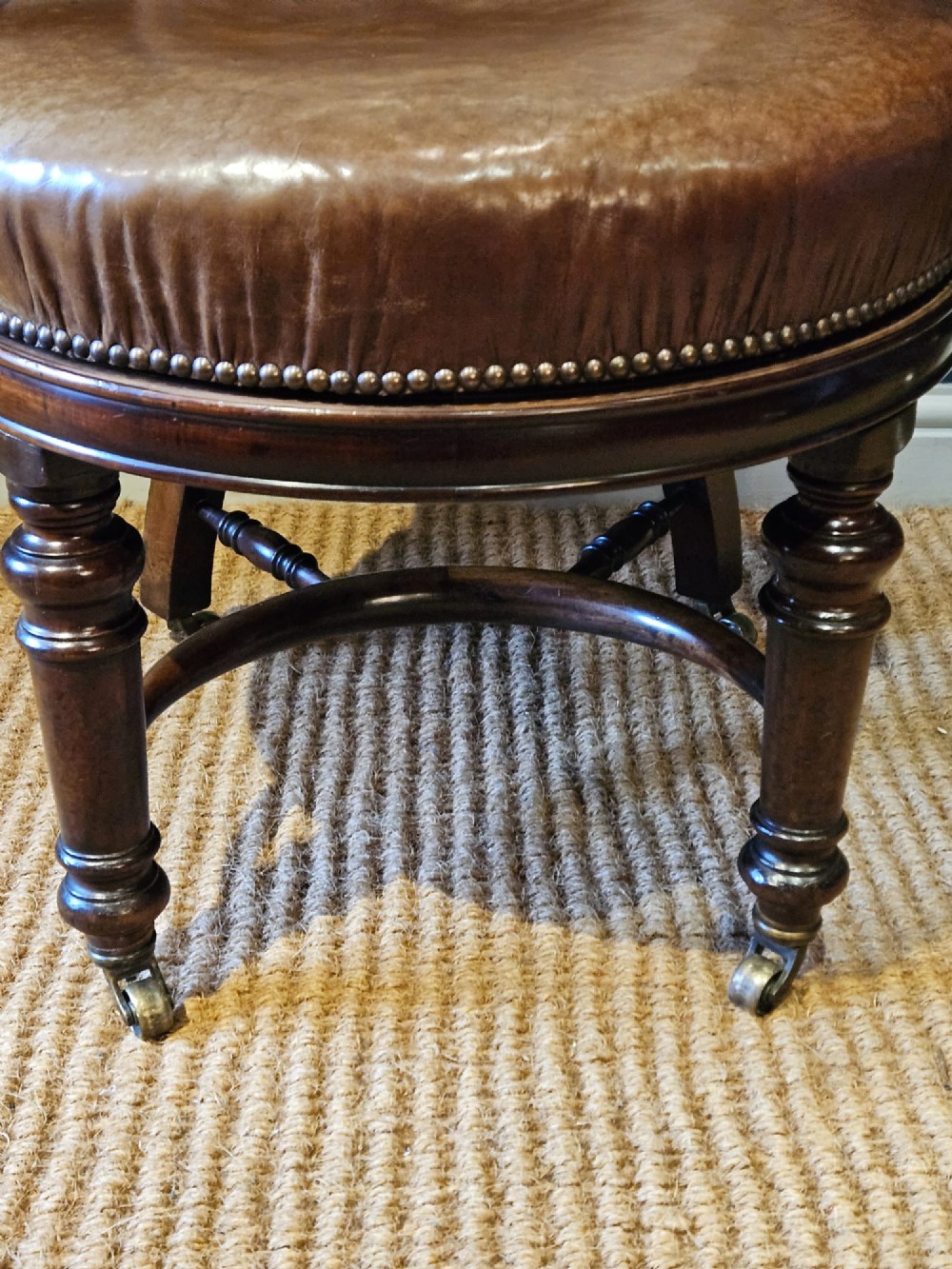 A Victorian revolving desk chair upholstered in tan hide leather with brass stud trim, curved armrests, and turned mahogany legs on brass castors. View V