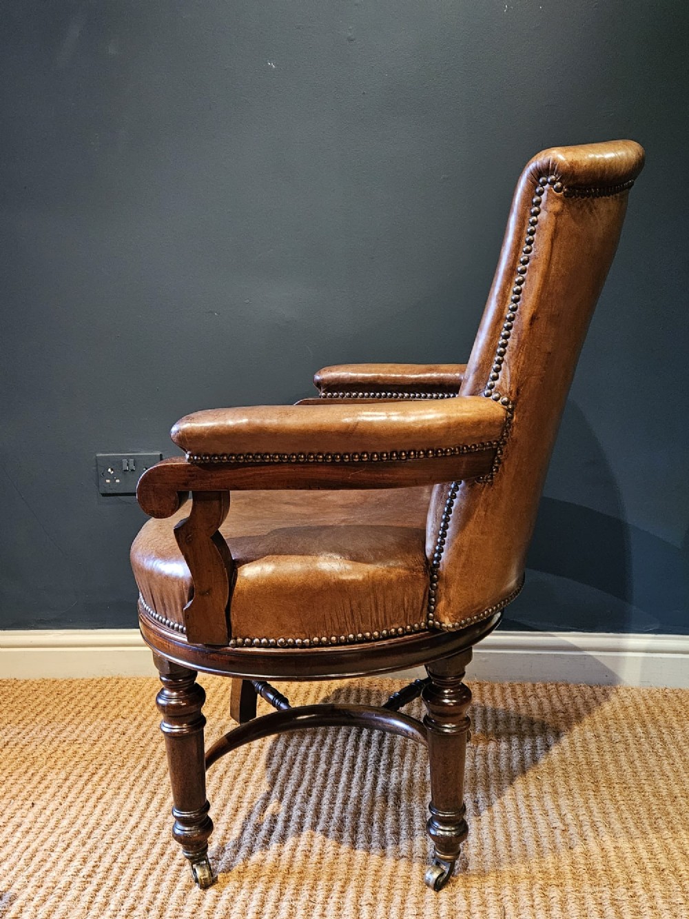 A Victorian revolving desk chair upholstered in tan hide leather with brass stud trim, curved armrests, and turned mahogany legs on brass castors. View VII