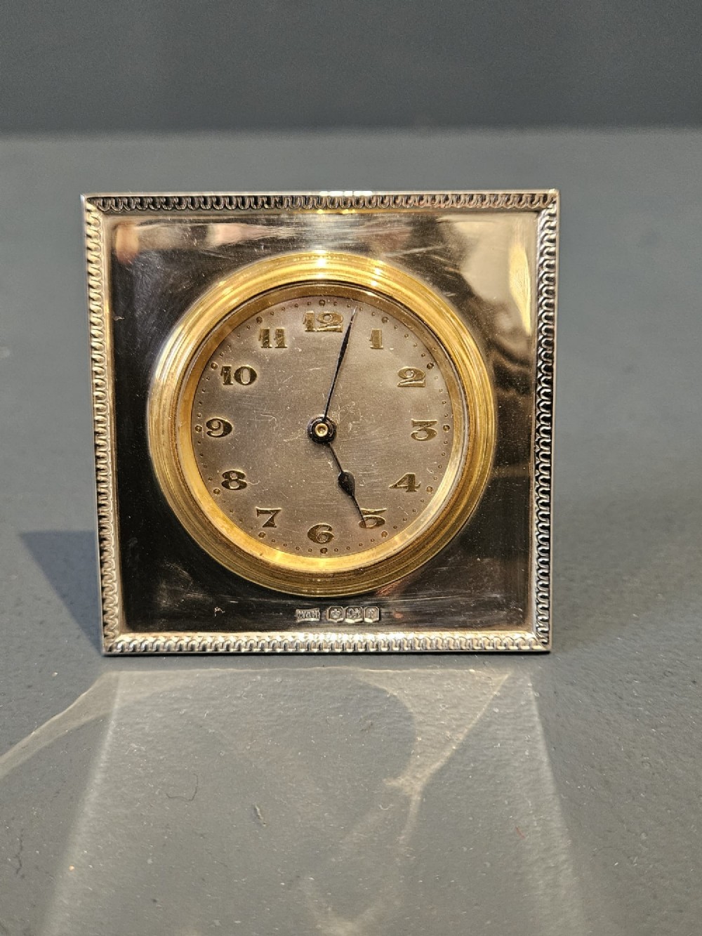 A square hallmarked sterling silver desk clock featuring a beaded outer frame, a brass inner bezel, and Arabic numerals on a metallic dial.