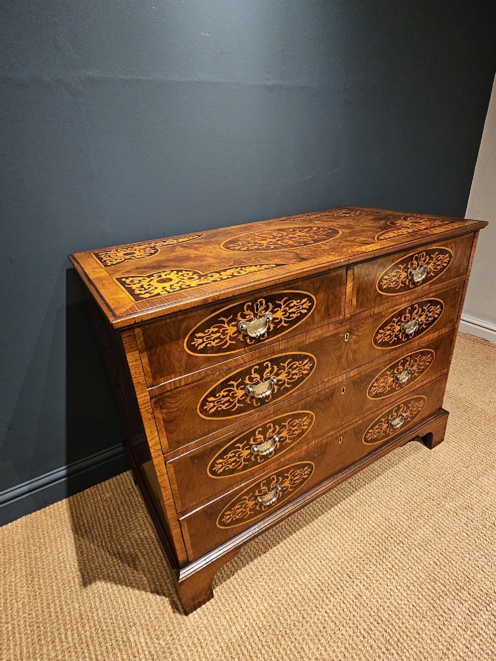 A George III period walnut chest of drawers featuring decorative oval marquetry inlays across four drawer fronts and brass handles, resting on bracket feet. View VII