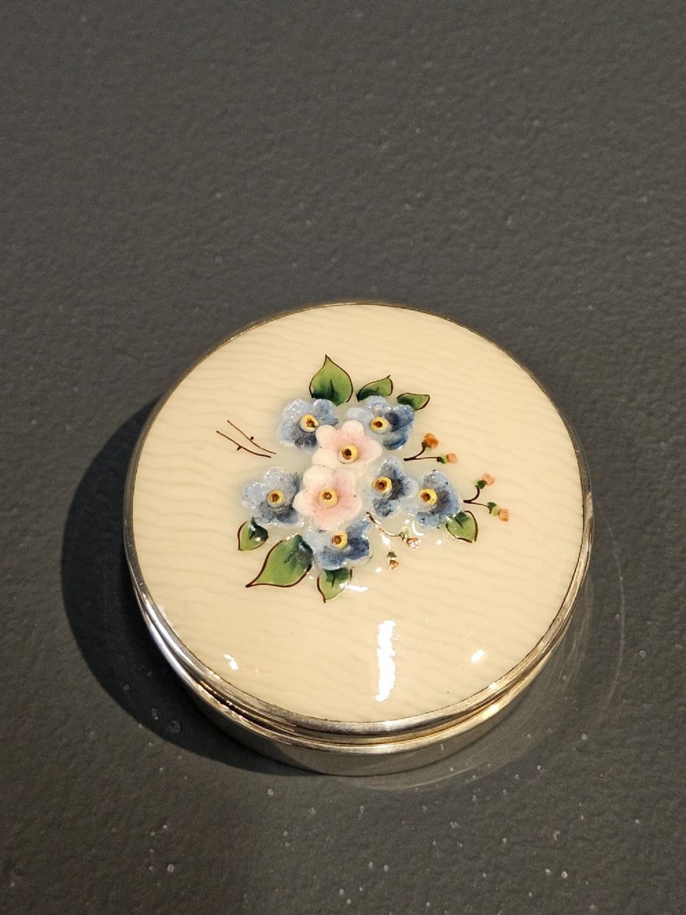 A small circular silver box featuring a cream-coloured porcelain lid decorated with hand-painted blue and pink flowers. View II