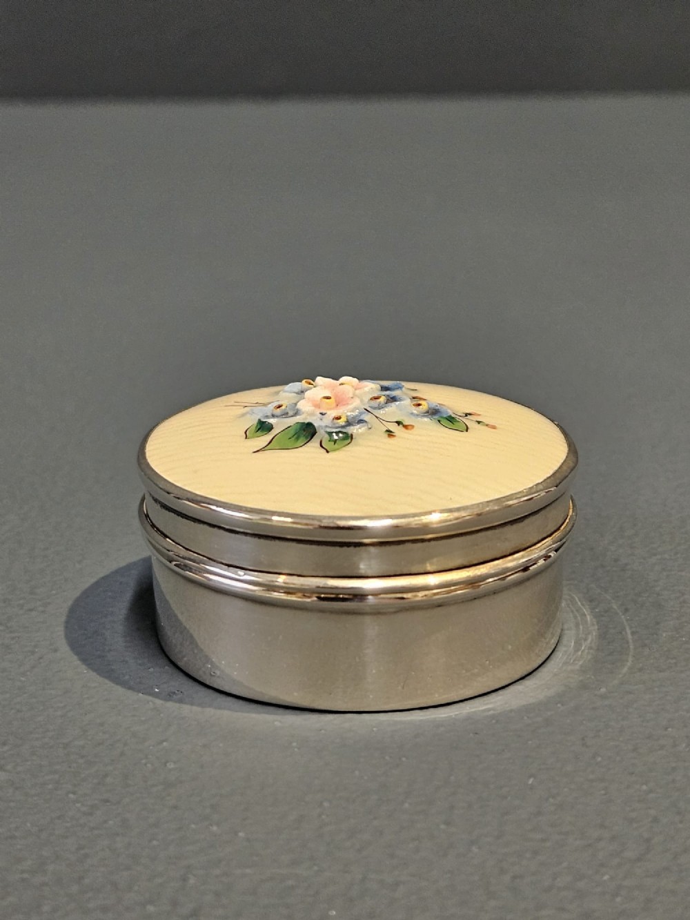 A small circular silver box featuring a cream-coloured porcelain lid decorated with hand-painted blue and pink flowers. View III