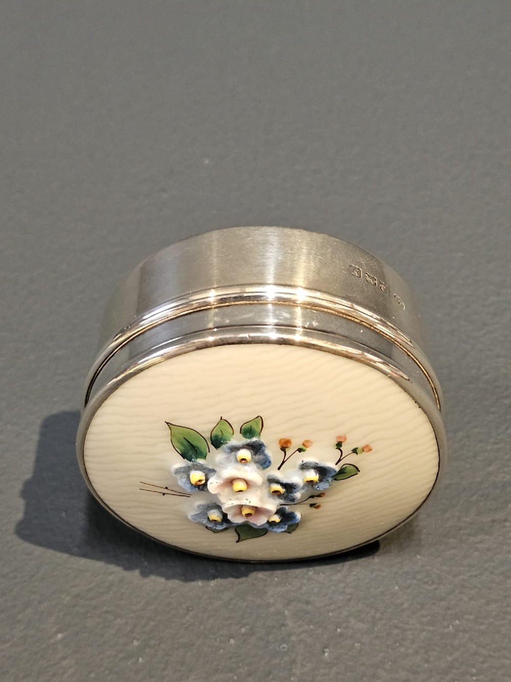 A small circular silver box featuring a cream-coloured porcelain lid decorated with hand-painted blue and pink flowers. View VI