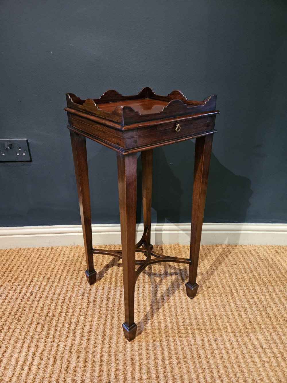 A tall Edwardian mahogany kettle stand featuring a scalloped gallery top, a small drawer with a brass pull, square tapered legs, and a crossed stretcher base.