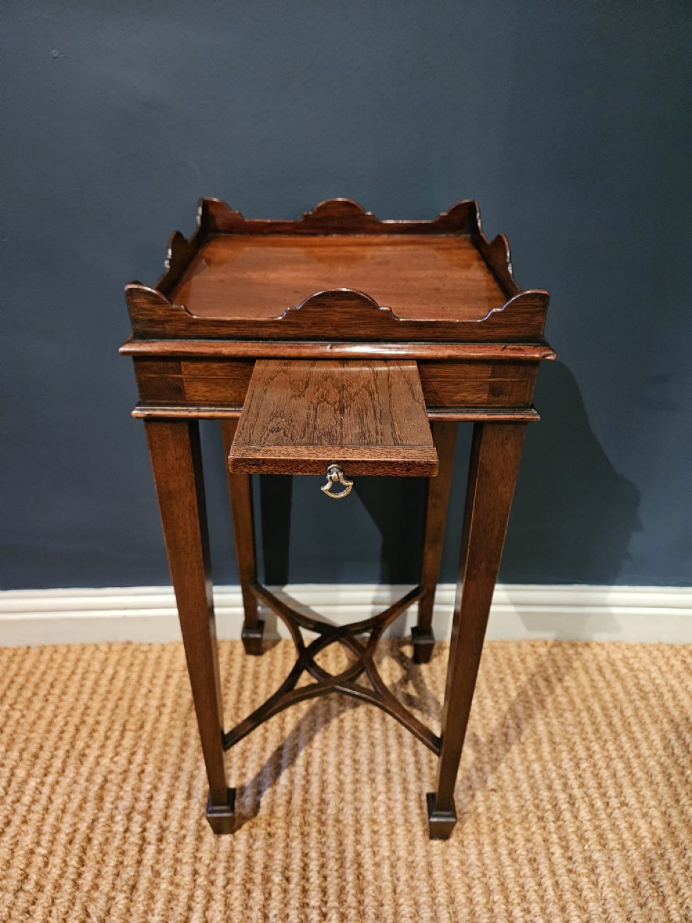 A tall Edwardian mahogany kettle stand featuring a scalloped gallery top, a small drawer with a brass pull, square tapered legs, and a crossed stretcher base. View II