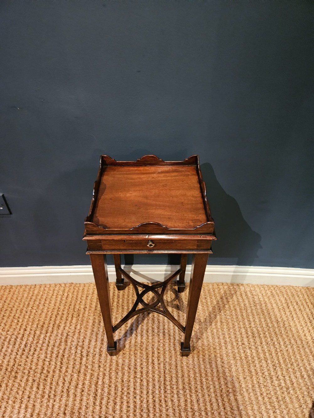 A tall Edwardian mahogany kettle stand featuring a scalloped gallery top, a small drawer with a brass pull, square tapered legs, and a crossed stretcher base. View III
