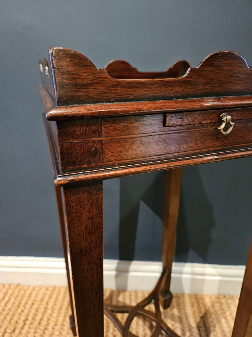 A tall Edwardian mahogany kettle stand featuring a scalloped gallery top, a small drawer with a brass pull, square tapered legs, and a crossed stretcher base. View IV