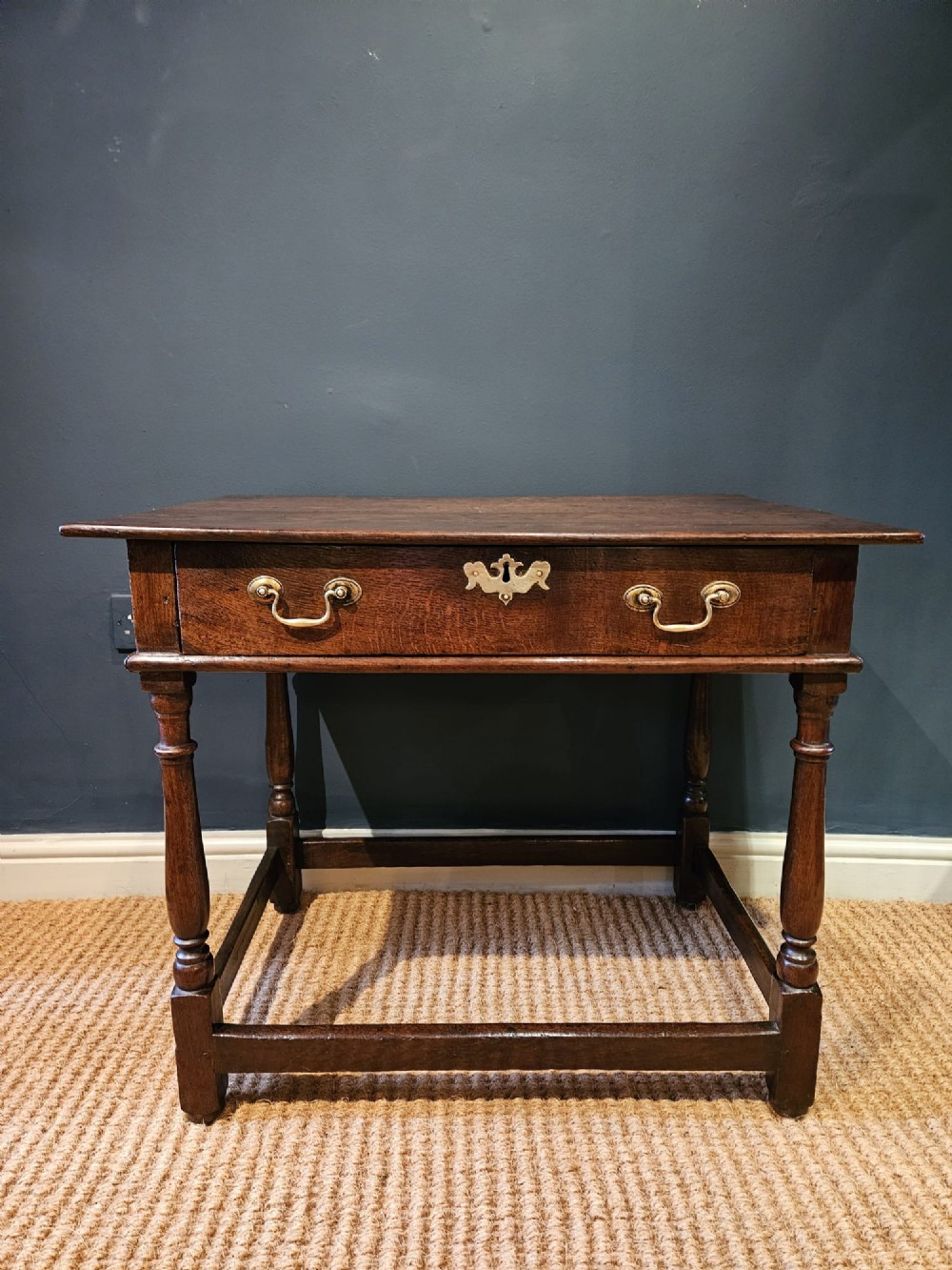 A dark oak George I side table featuring a single frieze drawer with brass drop handles and turned legs joined by stretchers. View IV