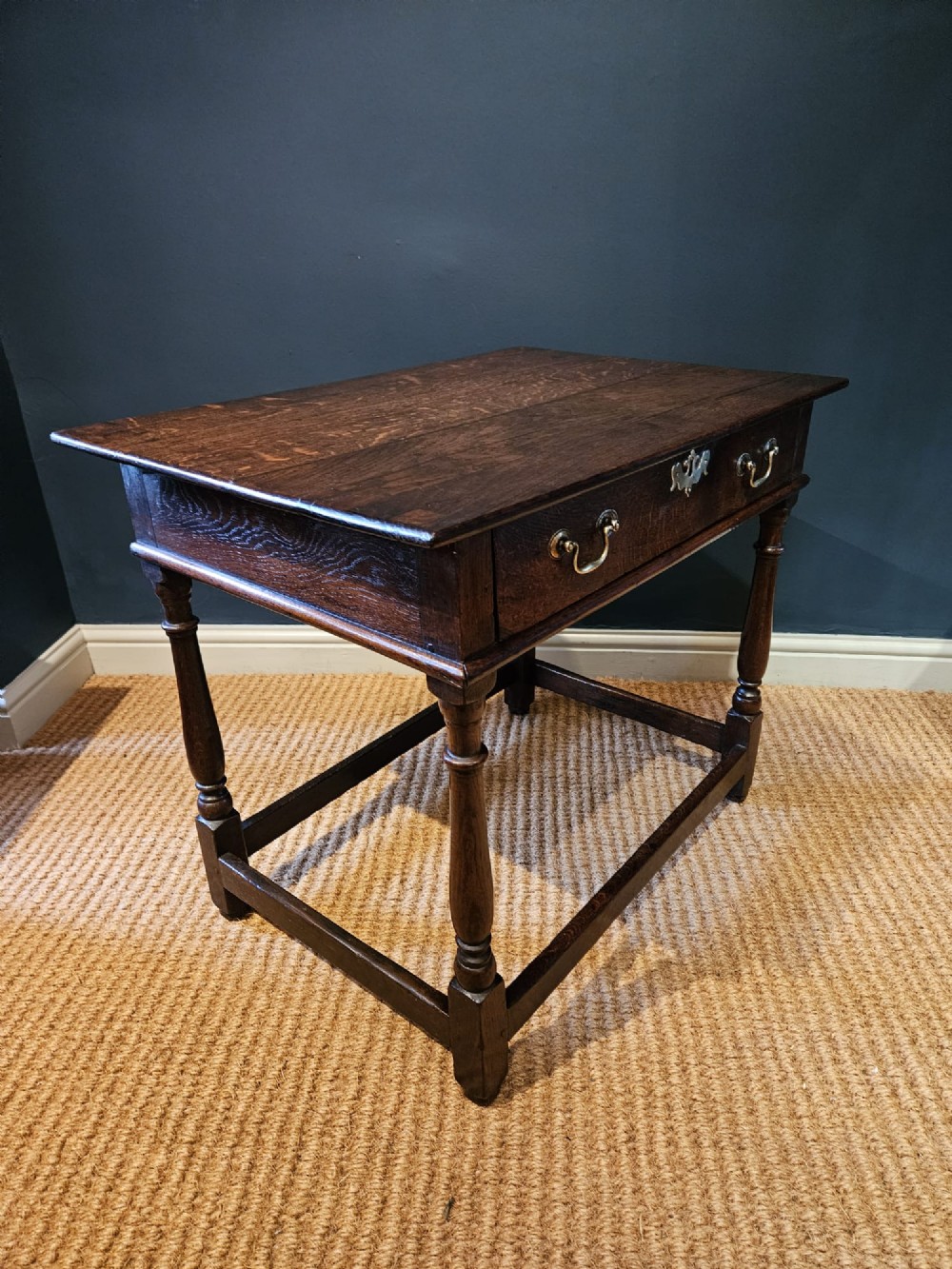 A dark oak George I side table featuring a single frieze drawer with brass drop handles and turned legs joined by stretchers. View V