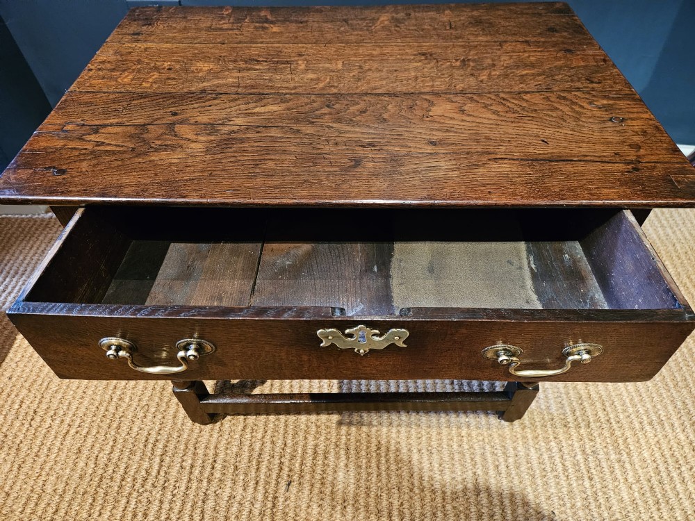 A dark oak George I side table featuring a single frieze drawer with brass drop handles and turned legs joined by stretchers. View VI