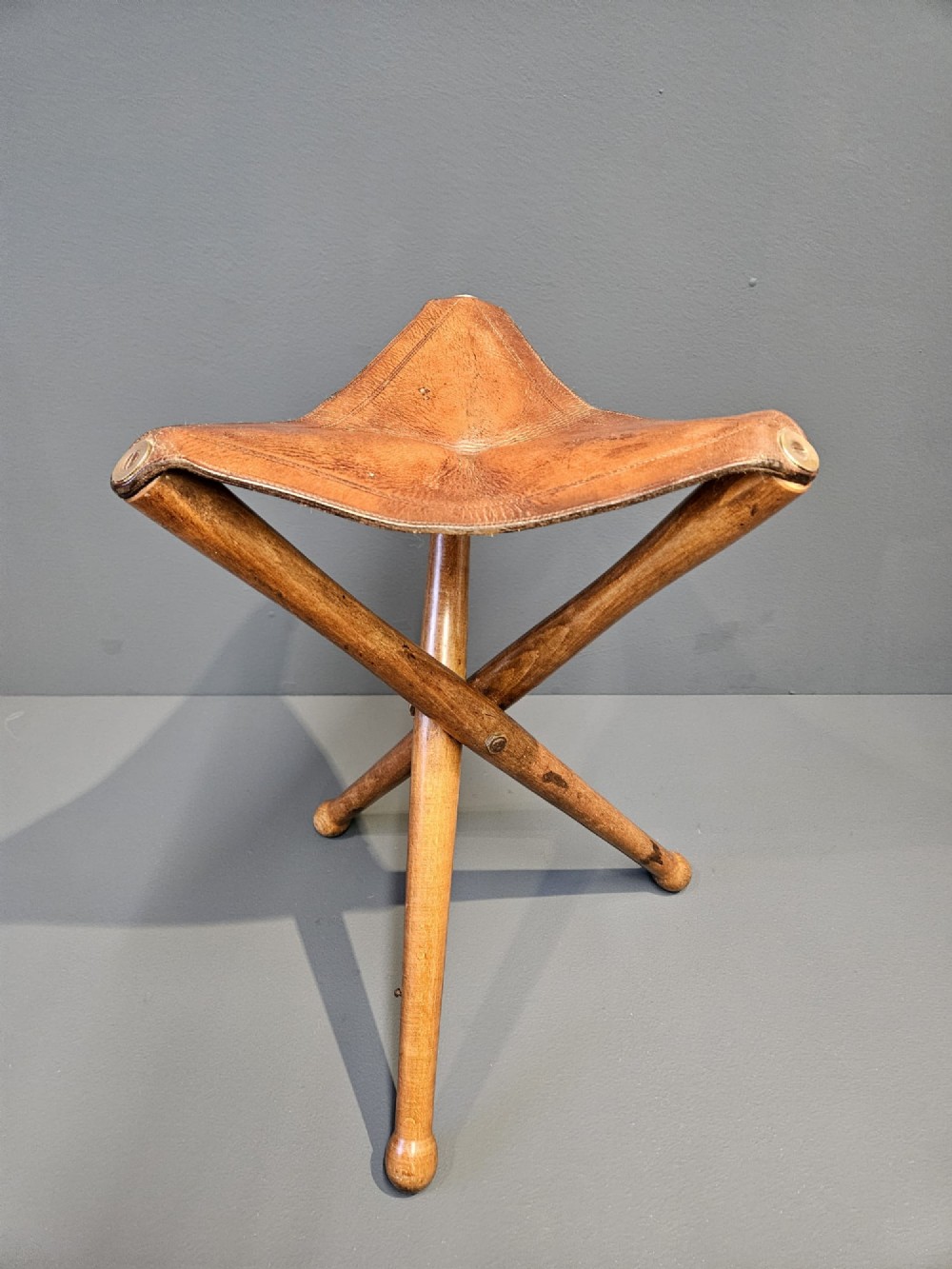 An Arts and Crafts period folding tripod campaign stool with three turned wooden legs and a stitched tan leather seat.