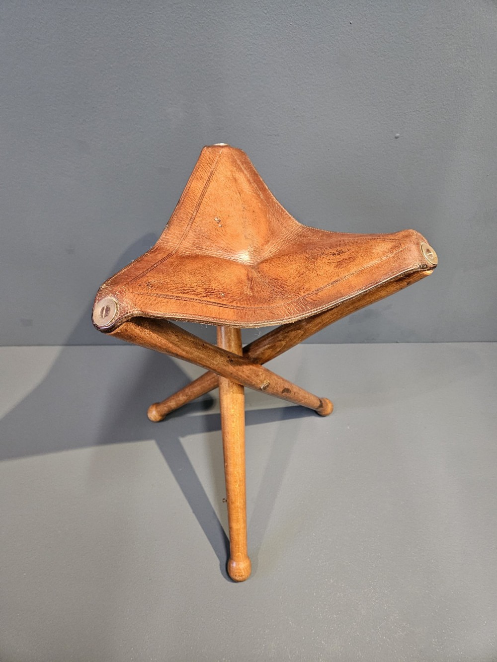 An Arts and Crafts period folding tripod campaign stool with three turned wooden legs and a stitched tan leather seat. View II