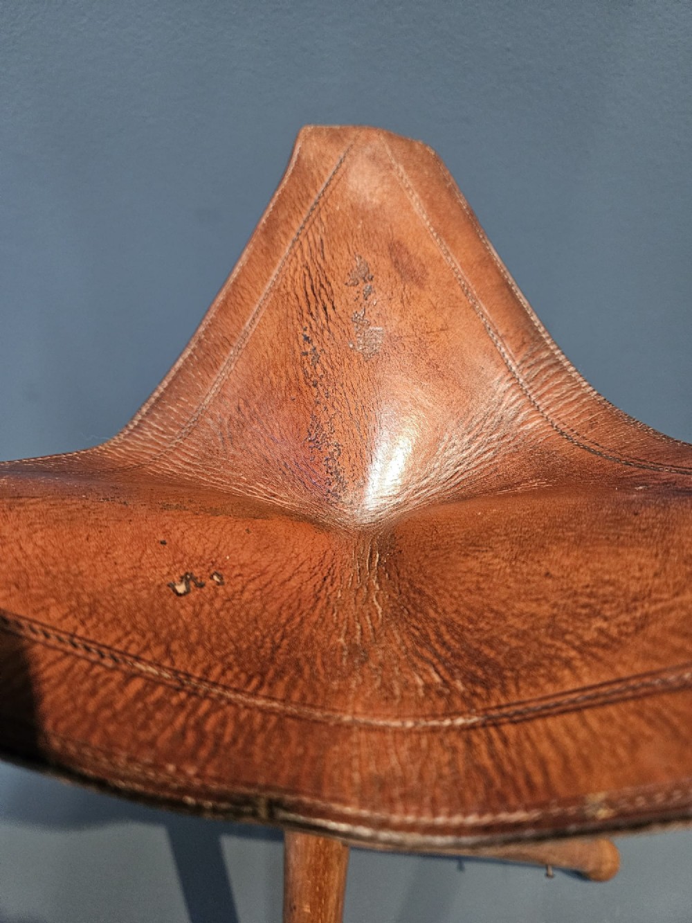 An Arts and Crafts period folding tripod campaign stool with three turned wooden legs and a stitched tan leather seat. View VI