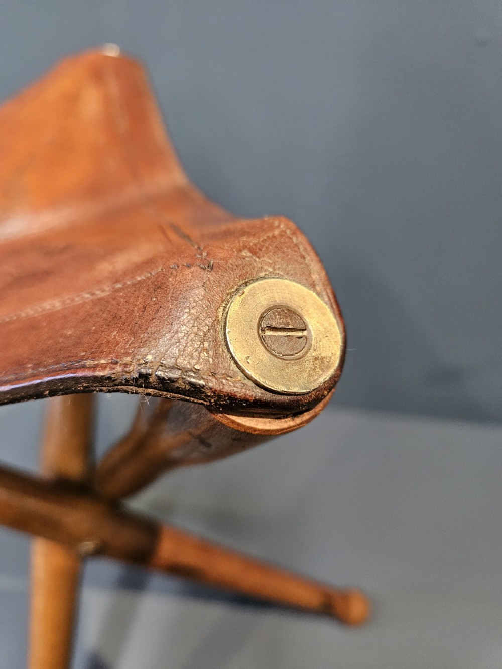 An Arts and Crafts period folding tripod campaign stool with three turned wooden legs and a stitched tan leather seat. View VIII