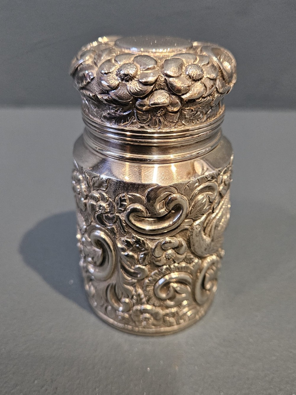 A cylindrical Victorian sterling silver cologne bottle with detailed chased floral scrollwork, bird motifs, and a matching screw cap. View II