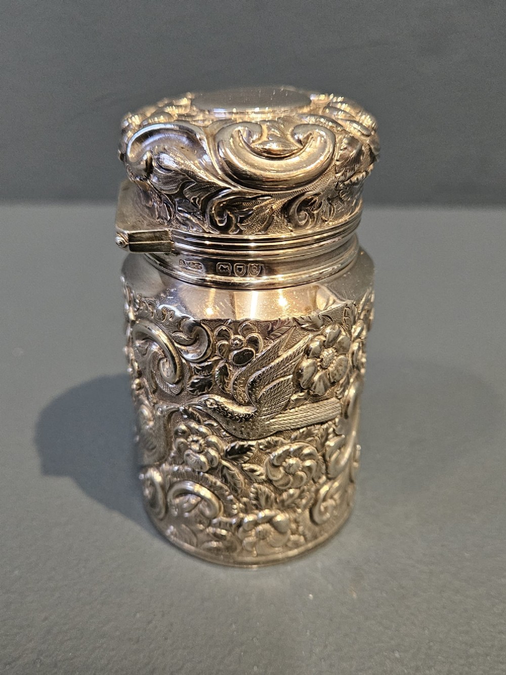 A cylindrical Victorian sterling silver cologne bottle with detailed chased floral scrollwork, bird motifs, and a matching screw cap. View IV