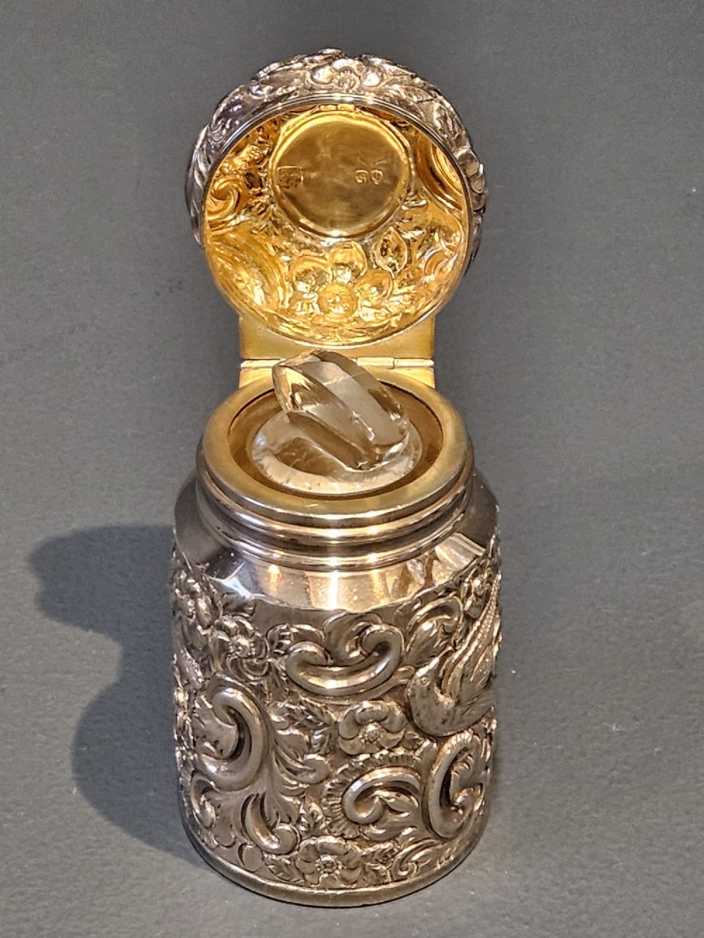 A cylindrical Victorian sterling silver cologne bottle with detailed chased floral scrollwork, bird motifs, and a matching screw cap. View V