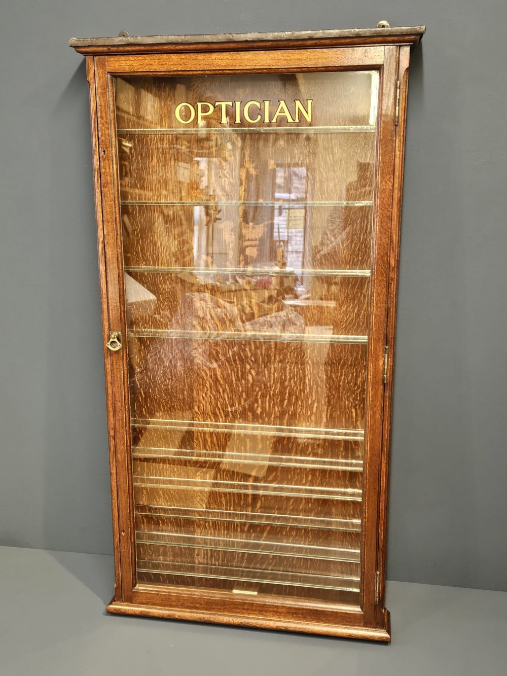 An Edwardian warm brown oak wall-mounted display cabinet with a single glass door featuring gold lettering that reads OPTICIAN over internal glass shelves.