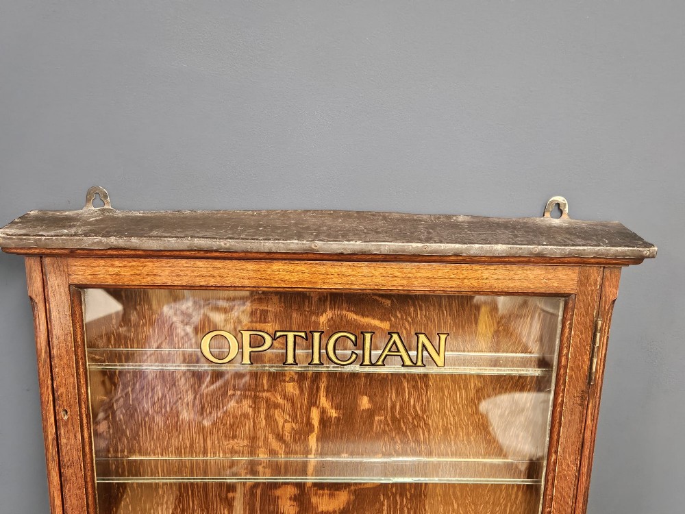 An Edwardian warm brown oak wall-mounted display cabinet with a single glass door featuring gold lettering that reads OPTICIAN over internal glass shelves. View II