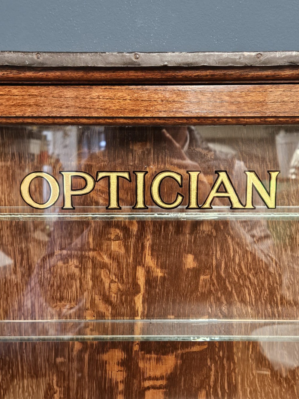 An Edwardian warm brown oak wall-mounted display cabinet with a single glass door featuring gold lettering that reads OPTICIAN over internal glass shelves. View III