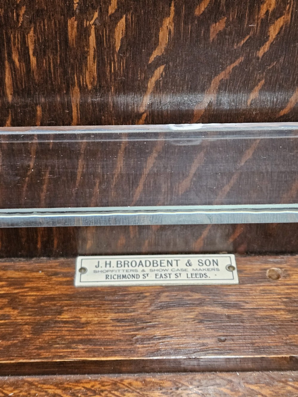 An Edwardian warm brown oak wall-mounted display cabinet with a single glass door featuring gold lettering that reads OPTICIAN over internal glass shelves. View VI