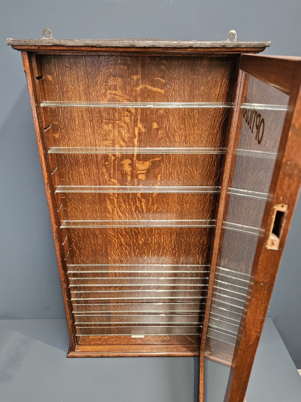 An Edwardian warm brown oak wall-mounted display cabinet with a single glass door featuring gold lettering that reads OPTICIAN over internal glass shelves. View VIII