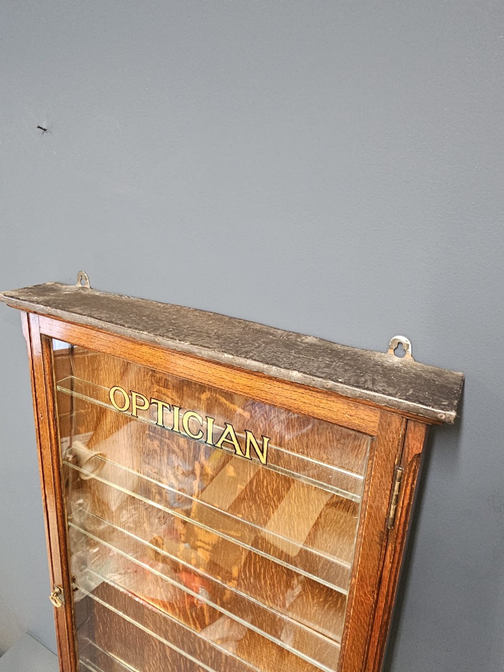 An Edwardian warm brown oak wall-mounted display cabinet with a single glass door featuring gold lettering that reads OPTICIAN over internal glass shelves. View IX