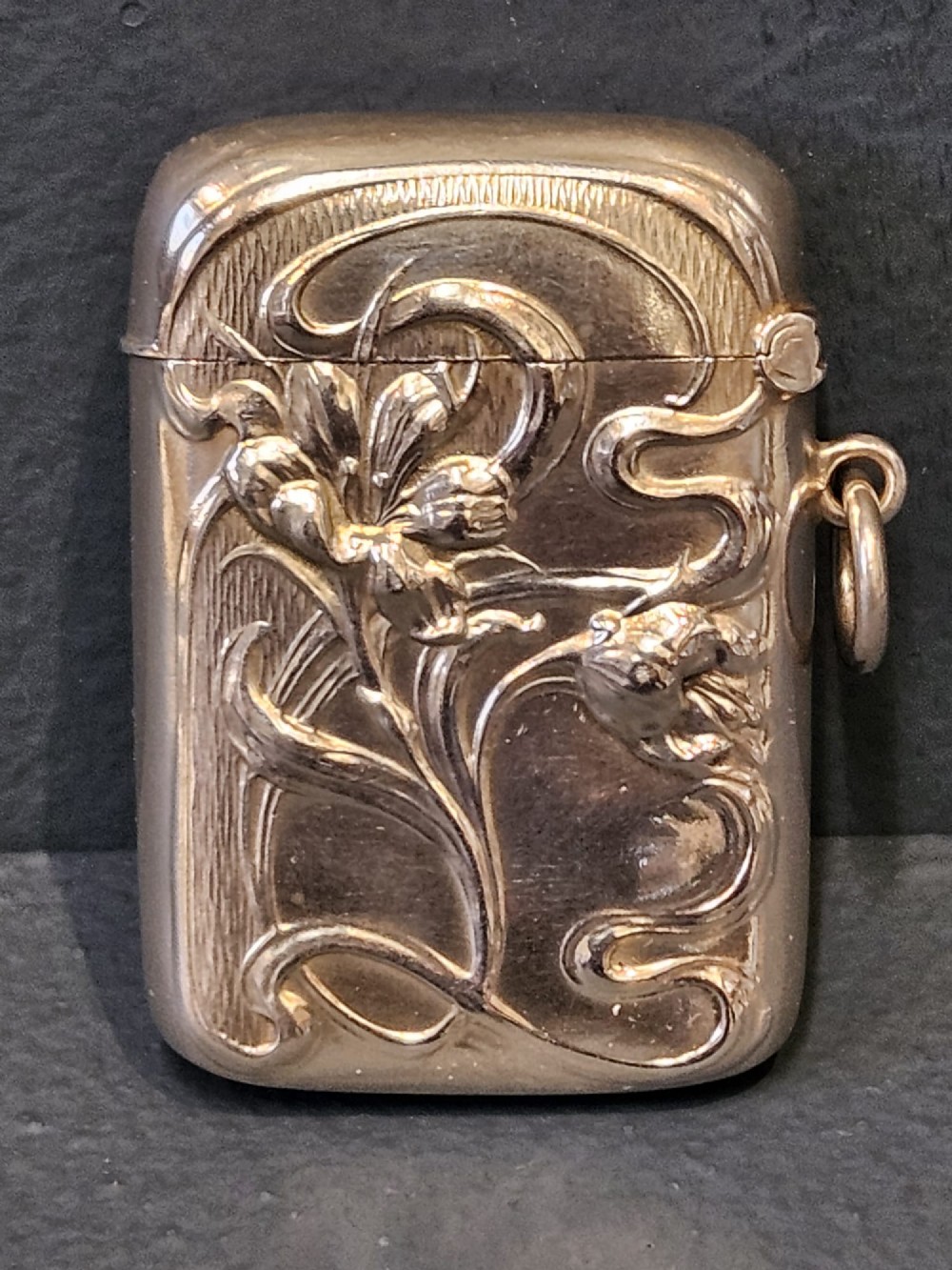 An Art Nouveau sterling silver vesta case featuring embossed floral motifs and whiplash scroll decoration with visible British hallmarks and a side suspension ring. View II