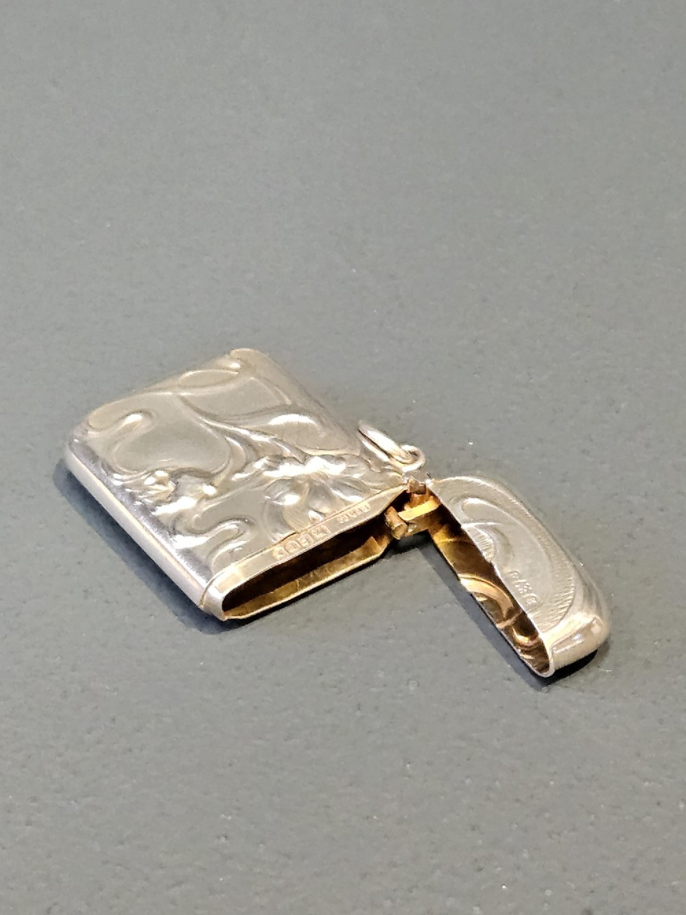 An Art Nouveau sterling silver vesta case featuring embossed floral motifs and whiplash scroll decoration with visible British hallmarks and a side suspension ring. View VI