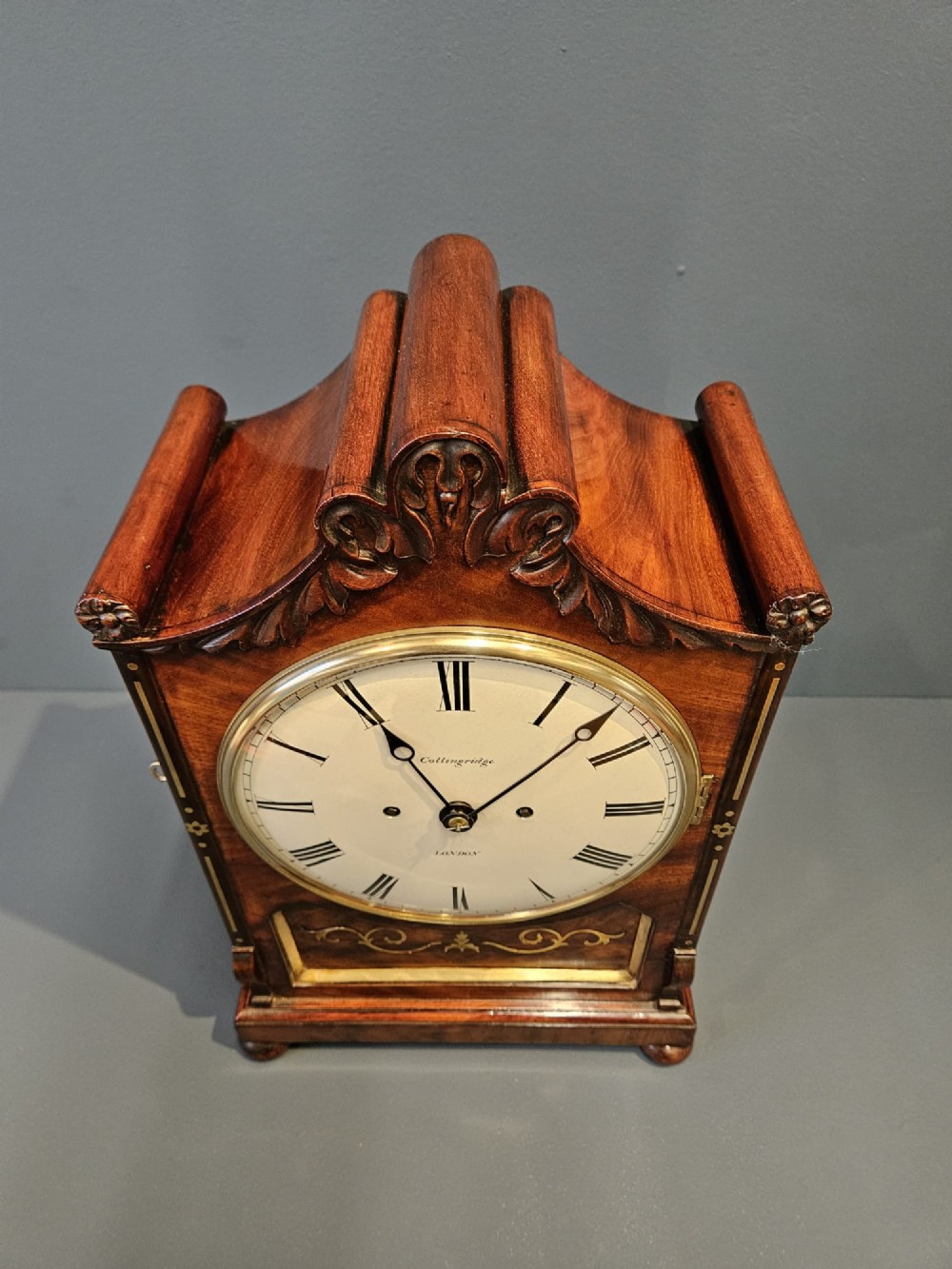 A William IV carved mahogany bracket clock featuring brass inlay decoration, a circular dial with Roman numerals, and a matching wall bracket. View VI