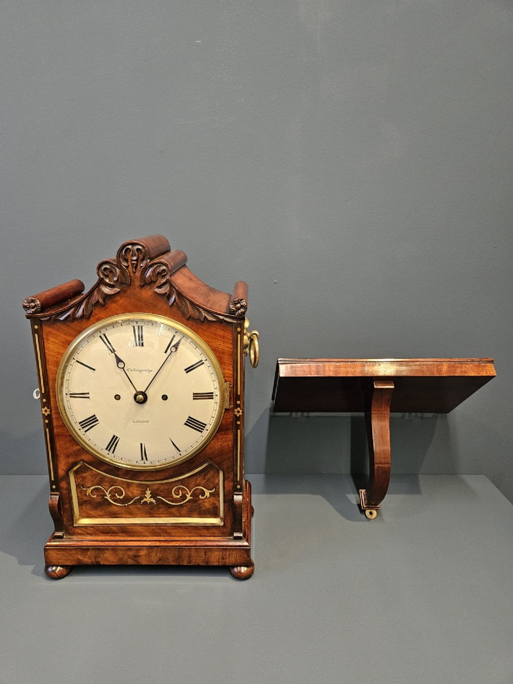 A William IV carved mahogany bracket clock featuring brass inlay decoration, a circular dial with Roman numerals, and a matching wall bracket. View X