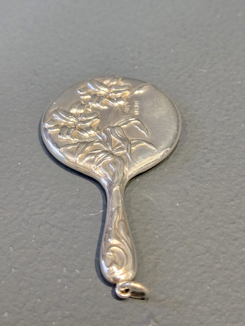 An Edwardian Art Nouveau sterling silver flirting mirror featuring embossed relief floral lily decoration and a suspension ring.