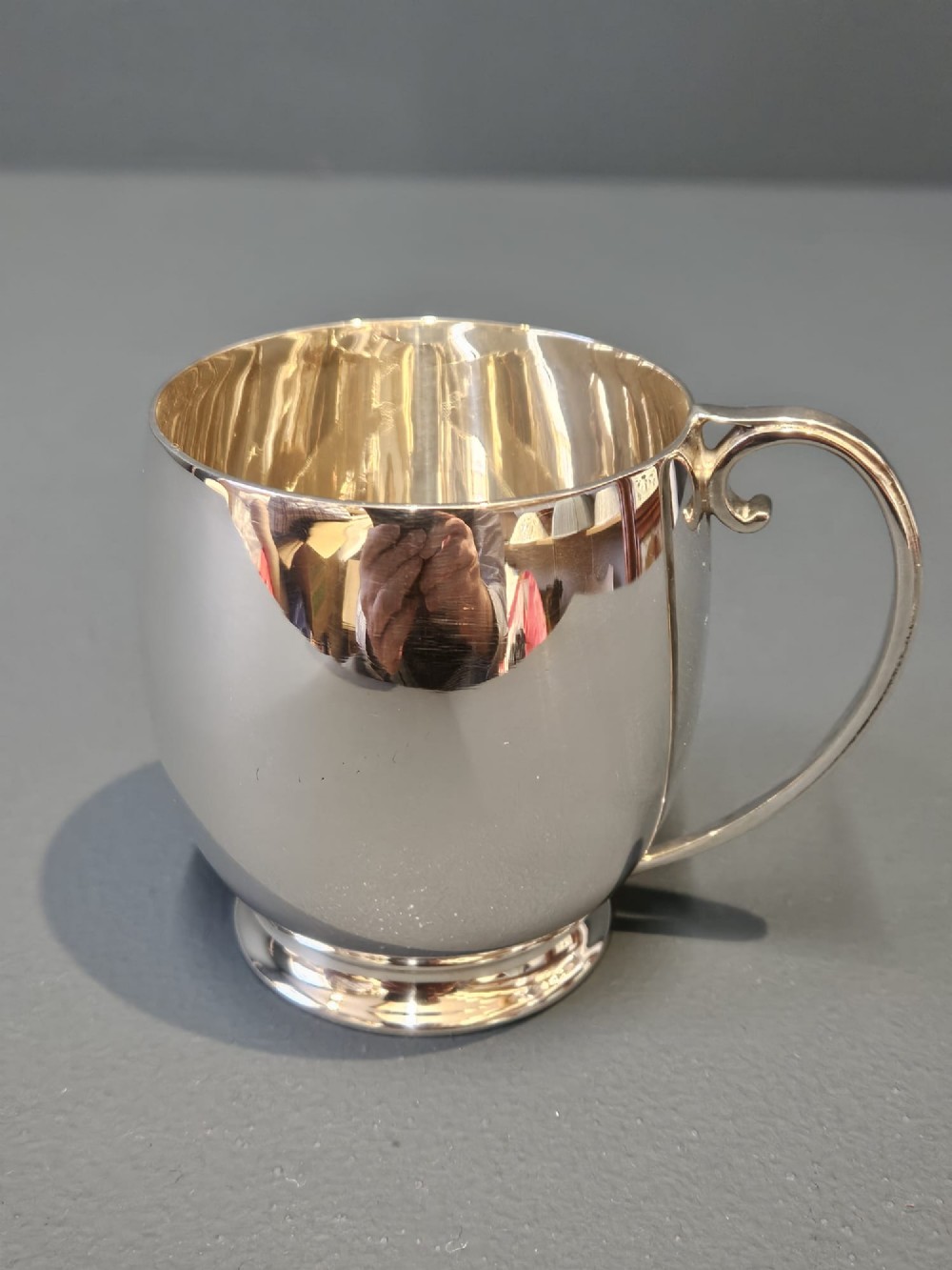 A polished silver mug featuring a bulbous body, a stepped base, and a scroll handle.