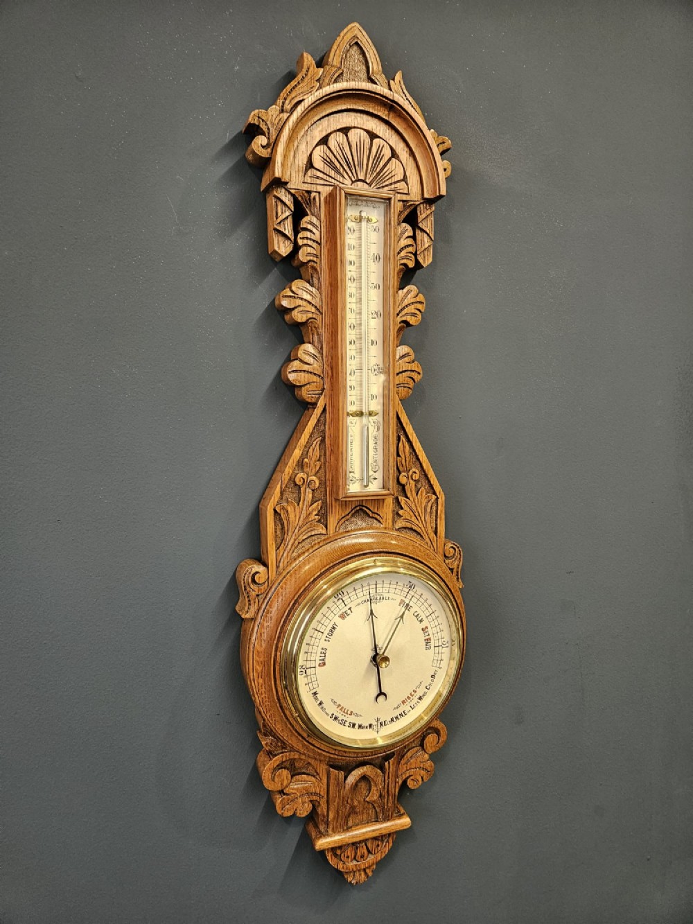 A carved Victorian golden oak banjo barometer with an integrated thermometer above a circular brass aneroid dial. View II