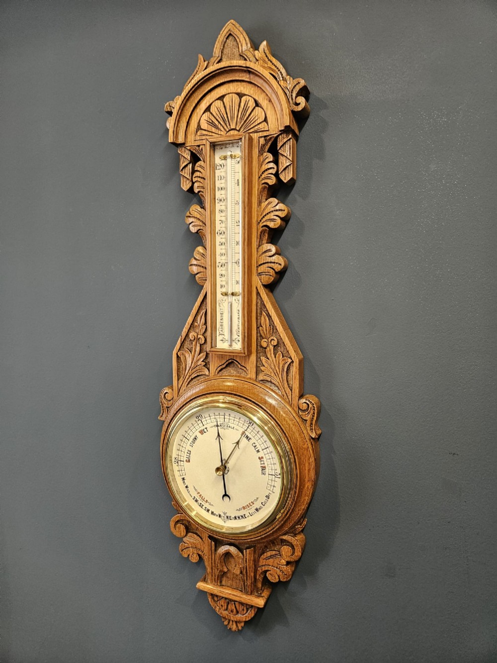 A carved Victorian golden oak banjo barometer with an integrated thermometer above a circular brass aneroid dial. View III