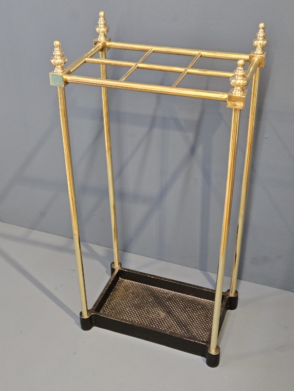 An Edwardian brass stick stand featuring four upright posts topped with urn finials, six upper compartments, and a textured black cast iron drip tray base.