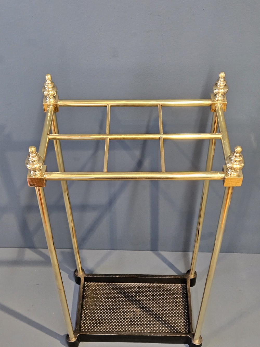 An Edwardian brass stick stand featuring four upright posts topped with urn finials, six upper compartments, and a textured black cast iron drip tray base. View II