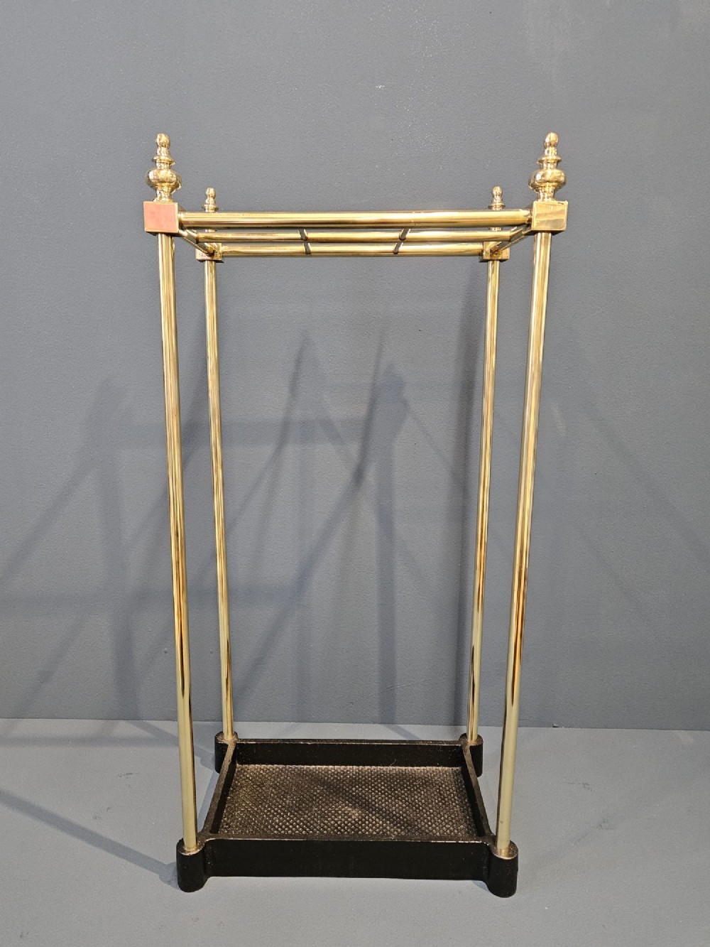 An Edwardian brass stick stand featuring four upright posts topped with urn finials, six upper compartments, and a textured black cast iron drip tray base. View IV