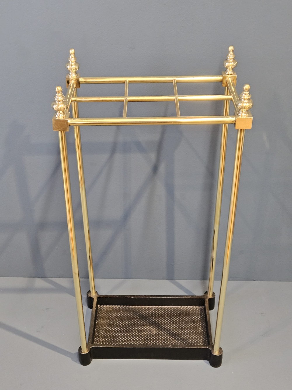 An Edwardian brass stick stand featuring four upright posts topped with urn finials, six upper compartments, and a textured black cast iron drip tray base. View VI