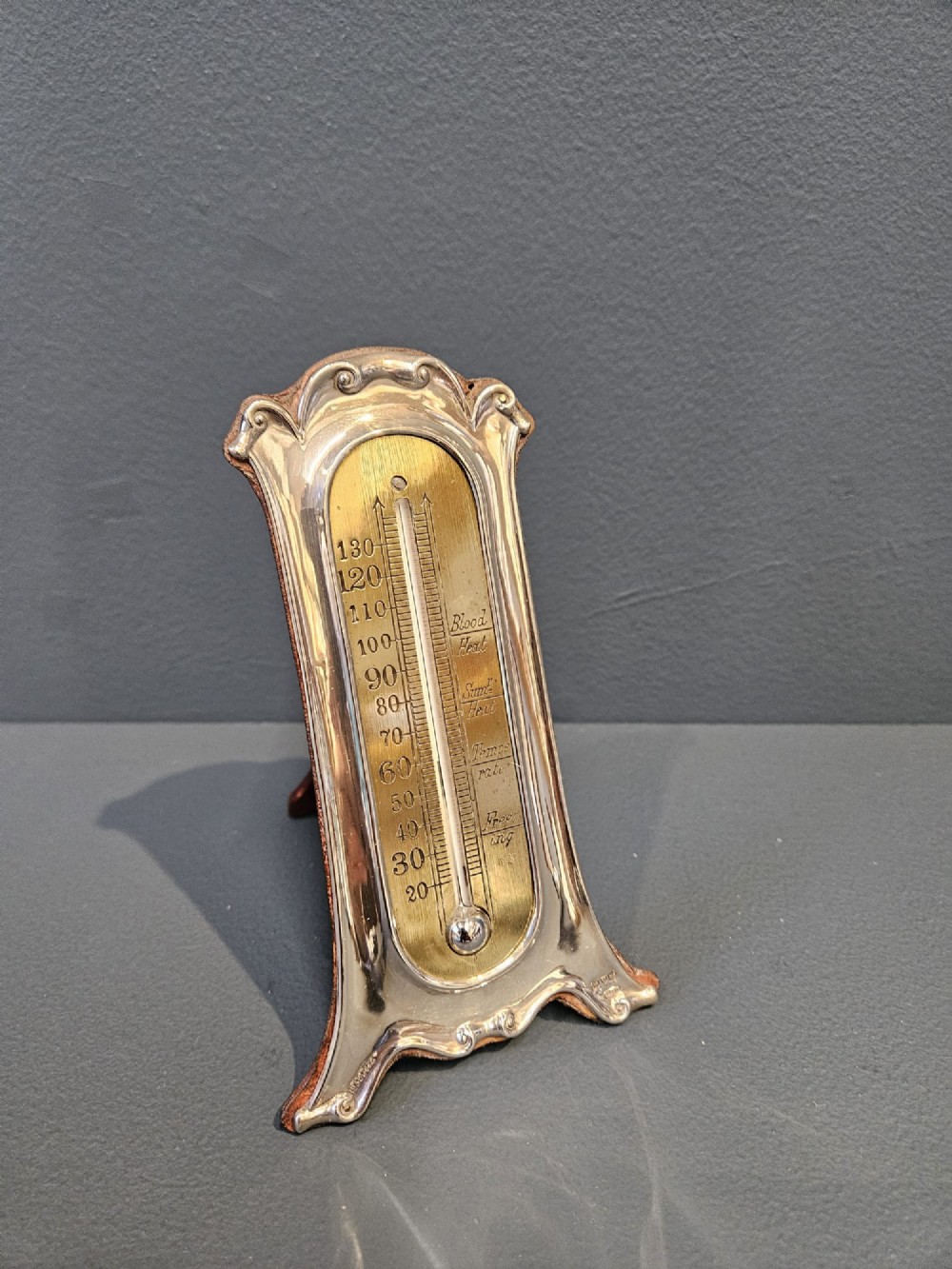 An Edwardian Art Nouveau silver desktop thermometer featuring sinuous repousse scrollwork, an engraved brass temperature dial, and a leather easel back.