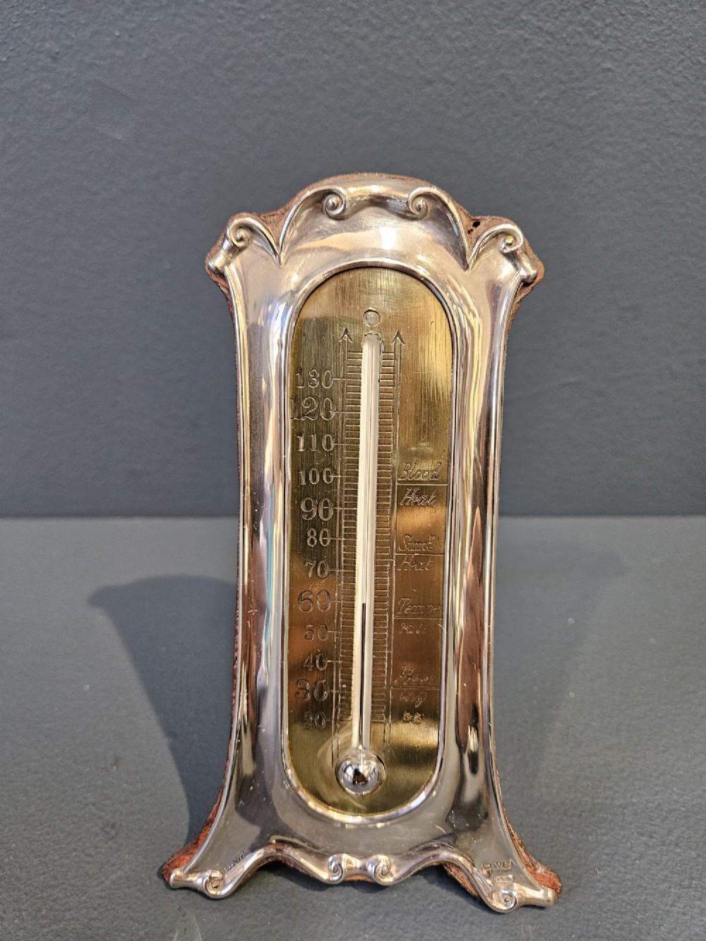 An Edwardian Art Nouveau silver desktop thermometer featuring sinuous repousse scrollwork, an engraved brass temperature dial, and a leather easel back. View III