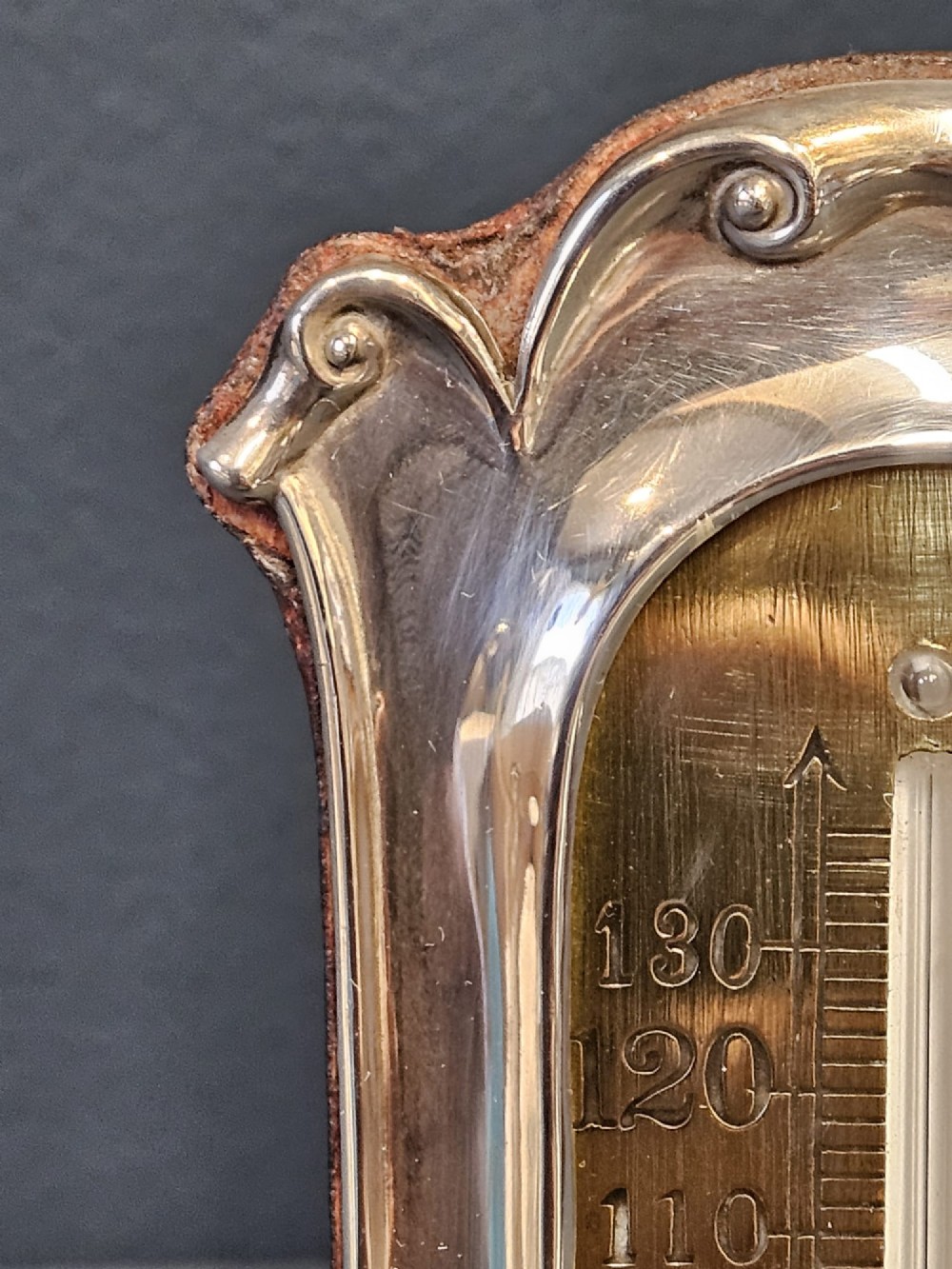 An Edwardian Art Nouveau silver desktop thermometer featuring sinuous repousse scrollwork, an engraved brass temperature dial, and a leather easel back. View IV