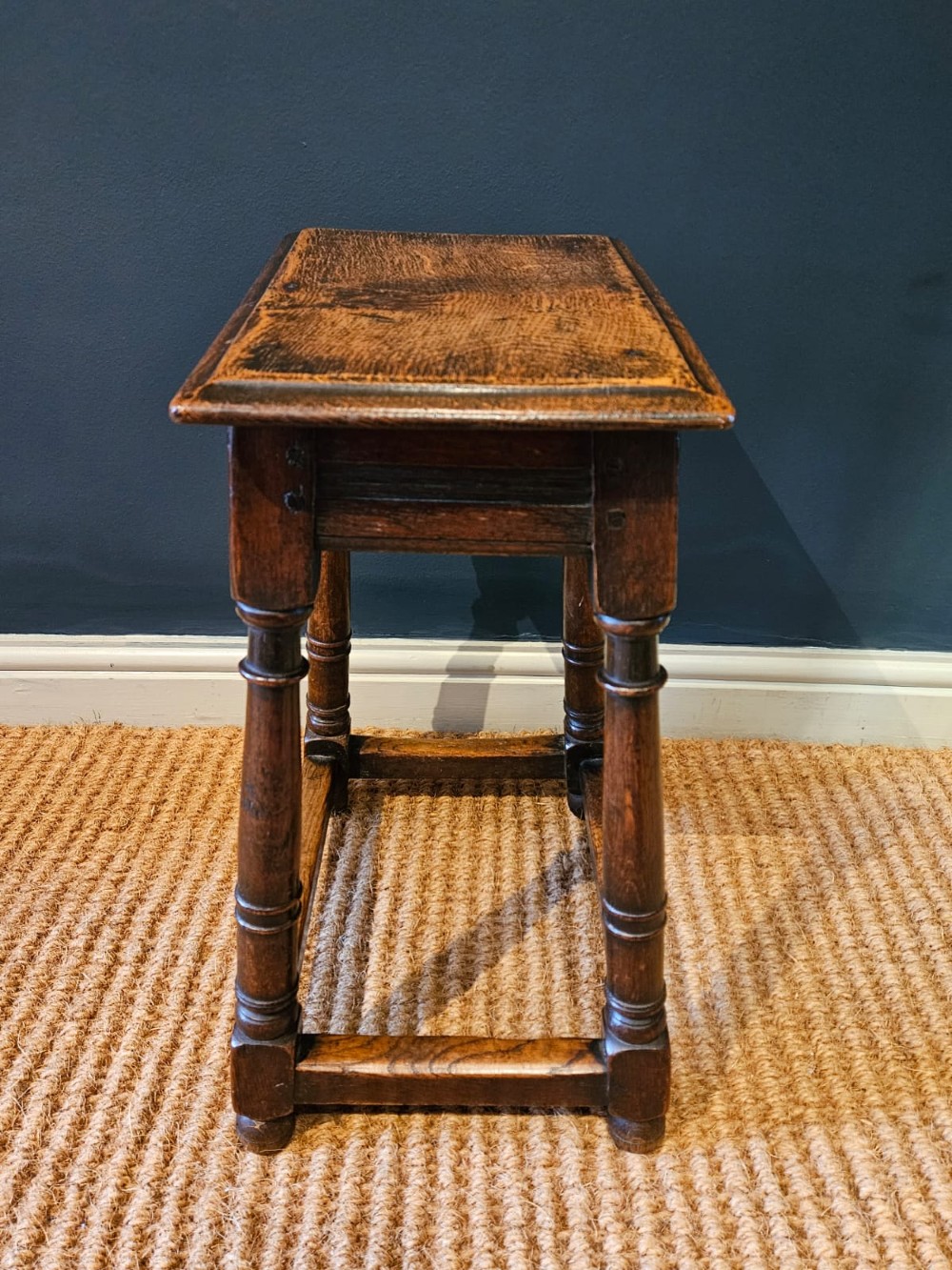 An antique dark oak joint stool featuring a rectangular top, moulded frieze, turned legs, and lower perimeter stretchers. View IV
