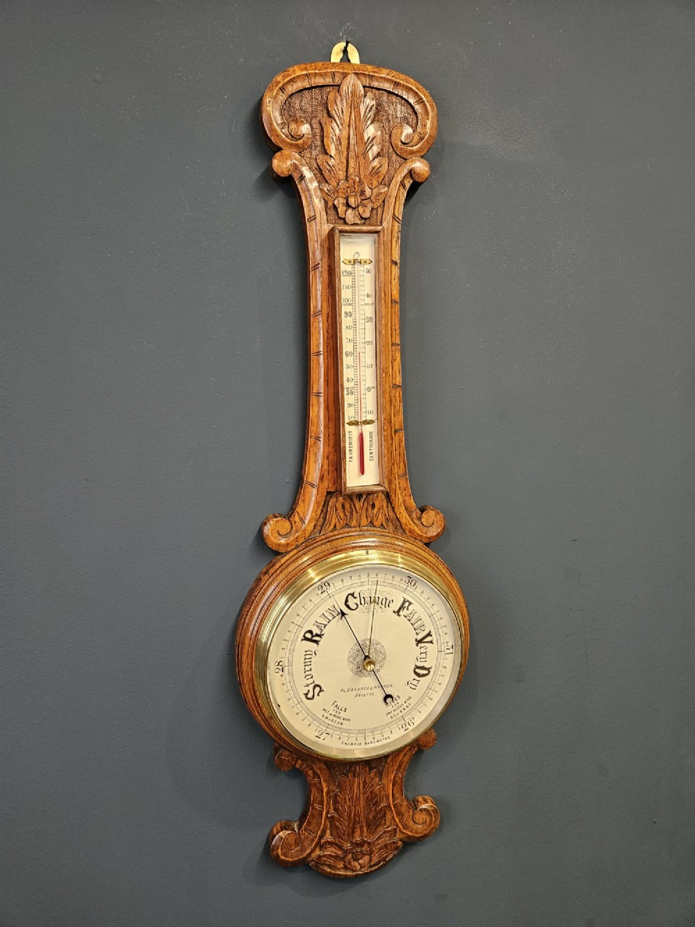 A Victorian carved oak banjo barometer featuring an upper thermometer tube and a lower circular brass-rimmed aneroid dial.