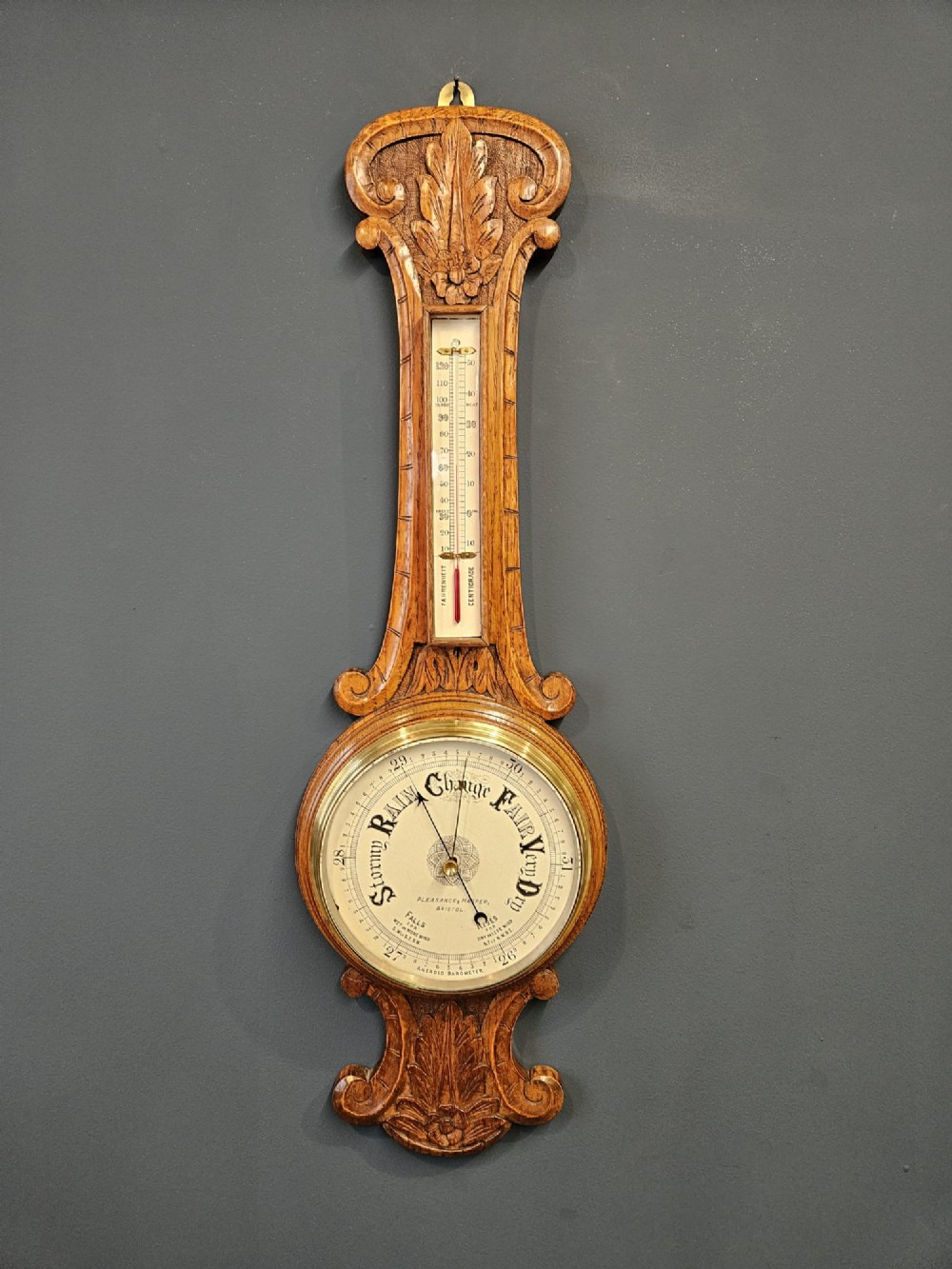 A Victorian carved oak banjo barometer featuring an upper thermometer tube and a lower circular brass-rimmed aneroid dial. View II